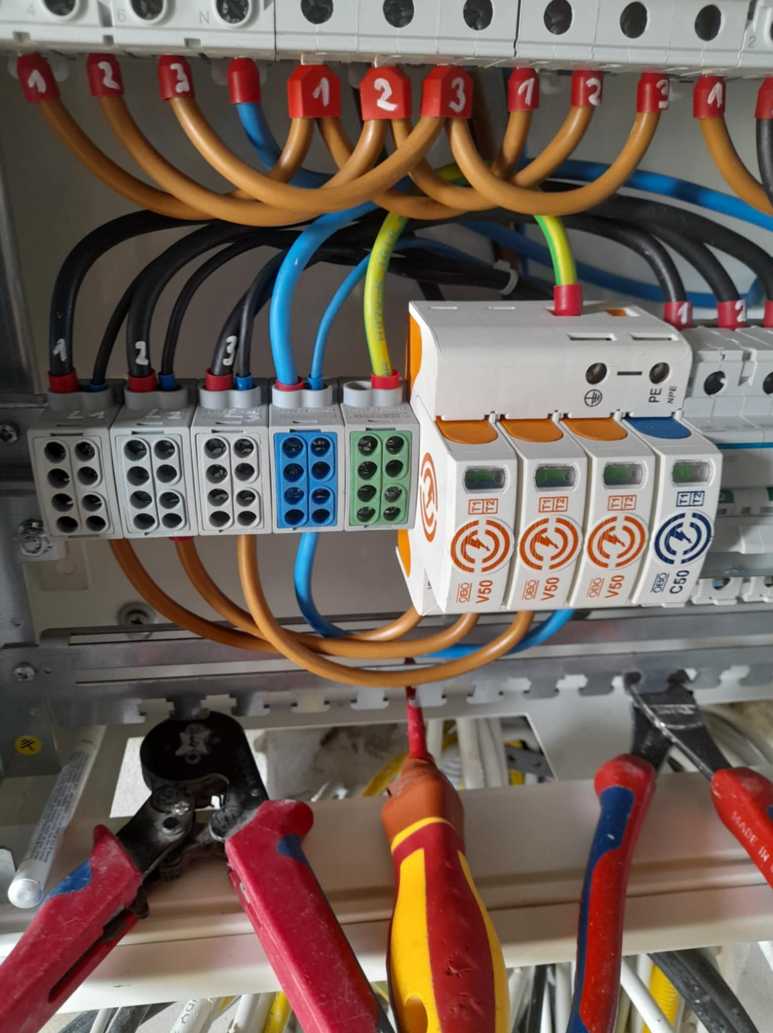 Rozdzielnica elektryczna z podłączonymi przewodami oznaczonymi numerami i zabezpieczeniami przepięciowymi. Widoczne narzędzia: kombinerki i śrubokręt.