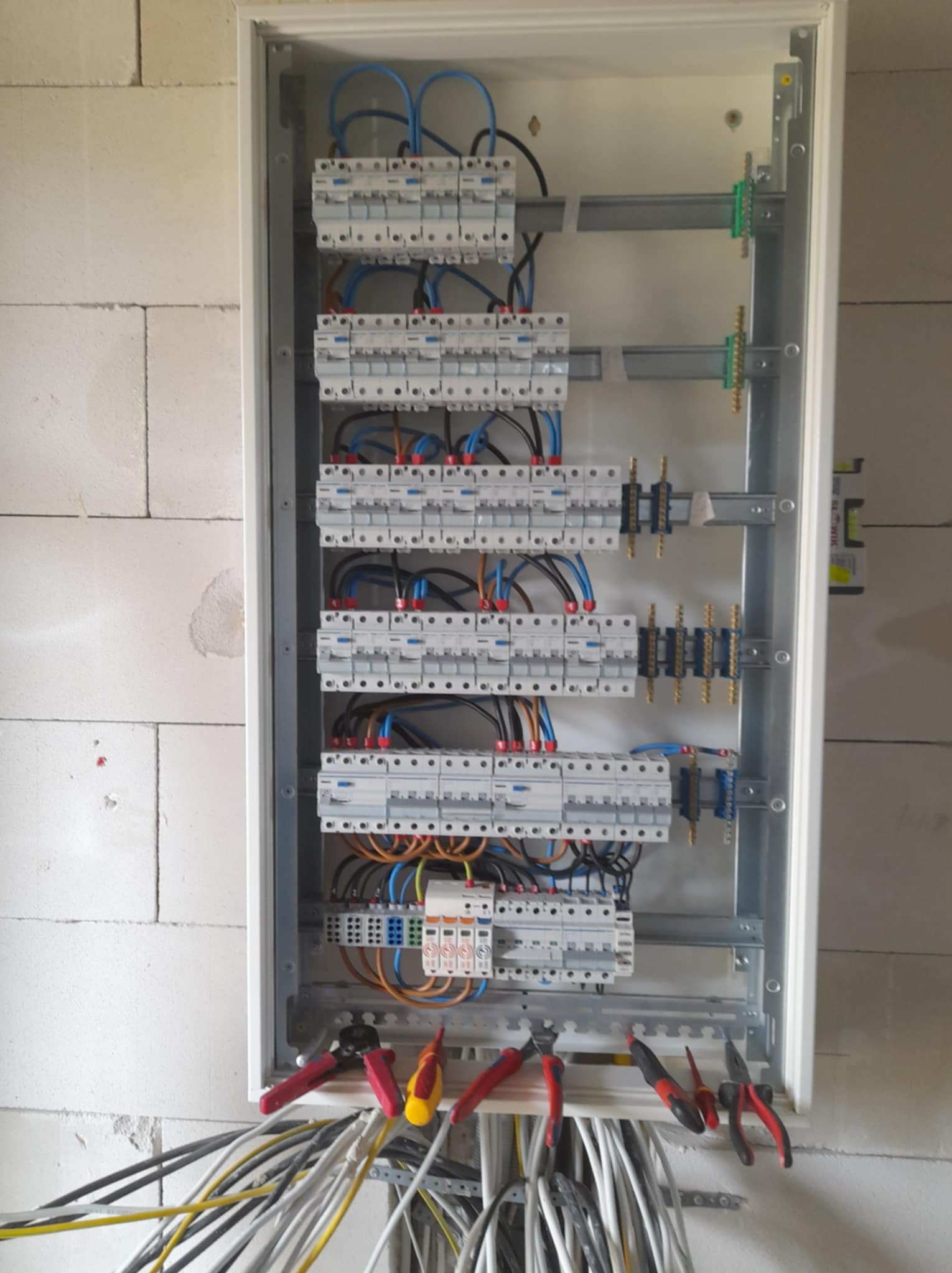 Rozdzielnia elektryczna w białej obudowie, z widocznymi bezpiecznikami, przewodami i narzędziami, zamontowana na ścianie z białych bloczków.