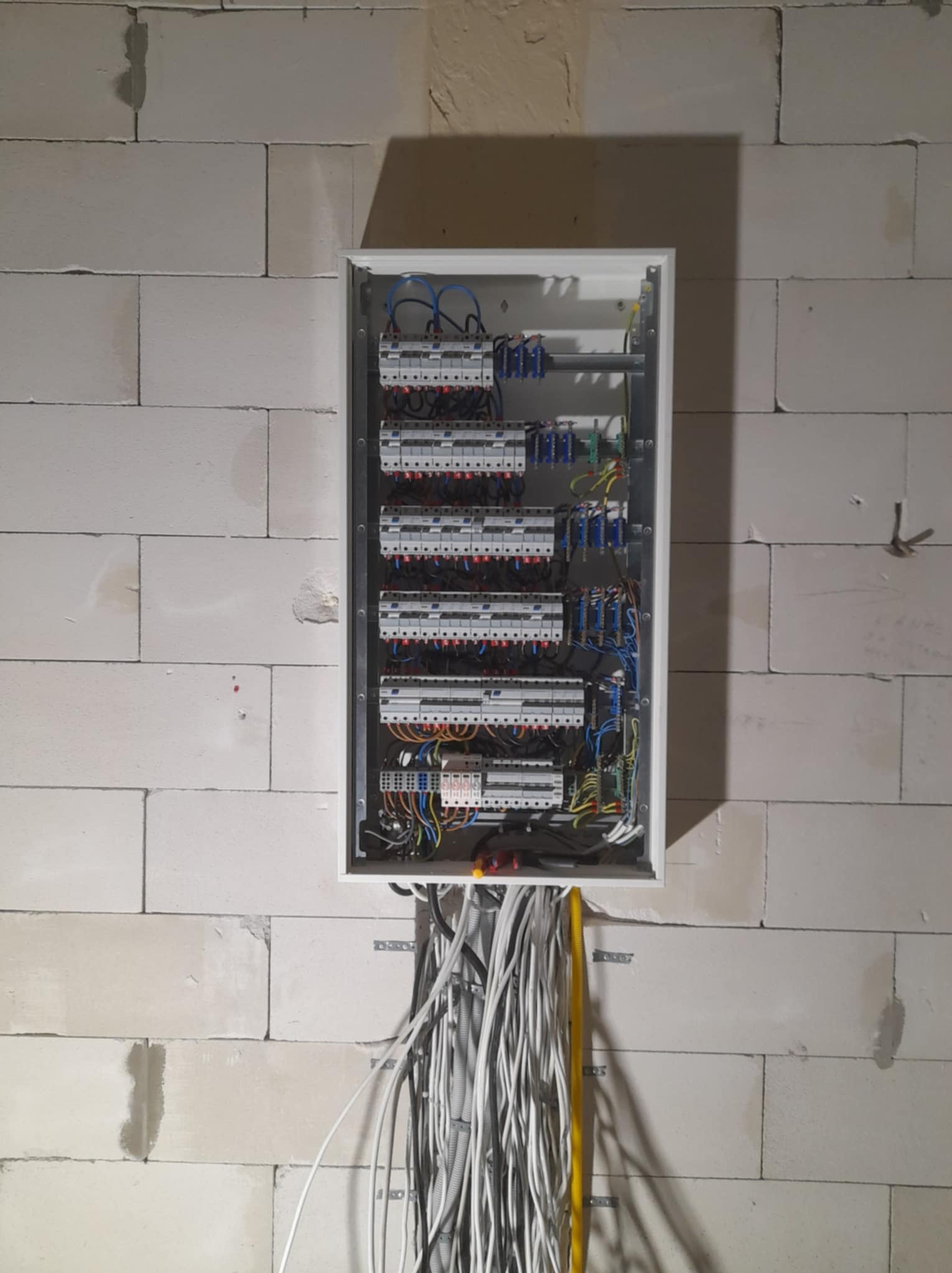 Rozdzielnica elektryczna z licznymi zabezpieczeniami, zamontowana na ścianie z betonu komórkowego. Widoczne kable wchodzące do skrzynki.