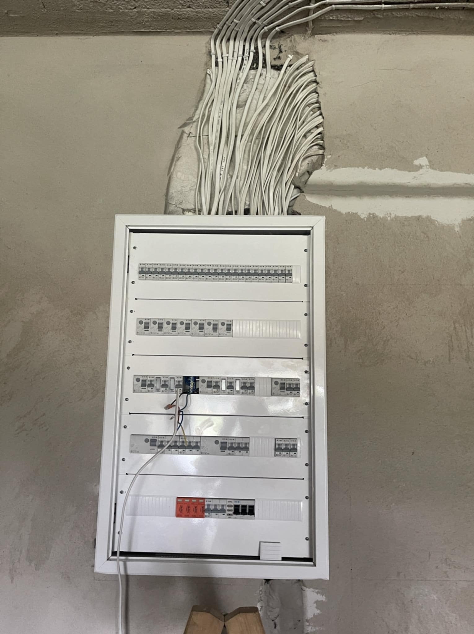 Rozdzielnica elektryczna w białej obudowie zamontowana na szarej ścianie, z widocznymi przewodami wchodzącymi do góry. Widoczne bezpieczniki i wyłączniki.