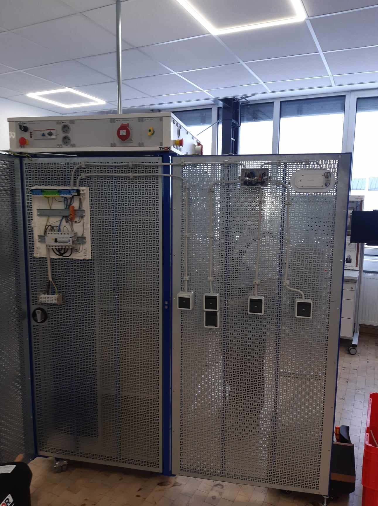 Tablica demonstracyjna z instalacją elektryczną i gniazdkami, umieszczona w jasnym pomieszczeniu z oknami i oświetleniem sufitowym.