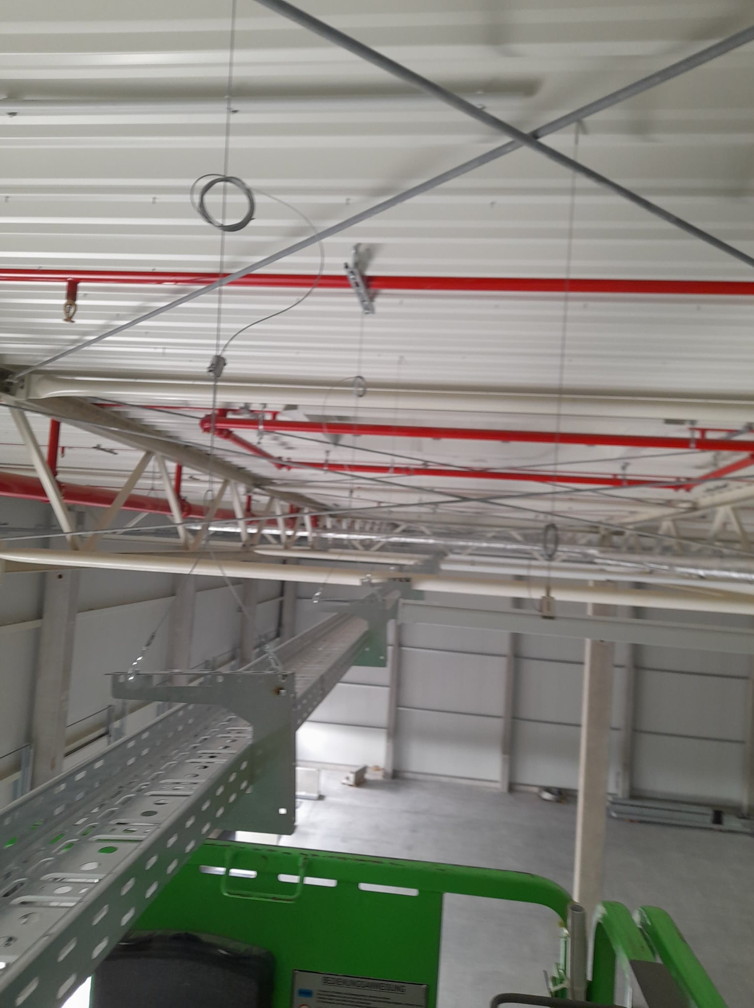 Szara trasa kablowa pod sufitem hali przemysłowej z widocznymi instalacjami. Ujęcie z podnośnika koszowego, widoczne elementy konstrukcji budynku.