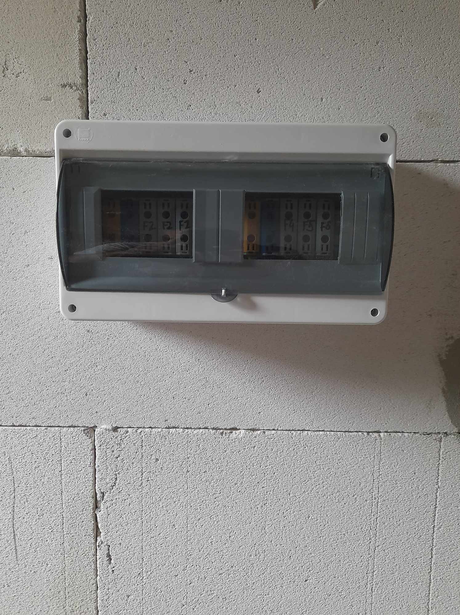 Szara skrzynka rozdzielcza z widocznymi bezpiecznikami i okablowaniem, zamontowana na surowej ścianie z betonu komórkowego. Widok z przodu, oświetlenie dzienne.