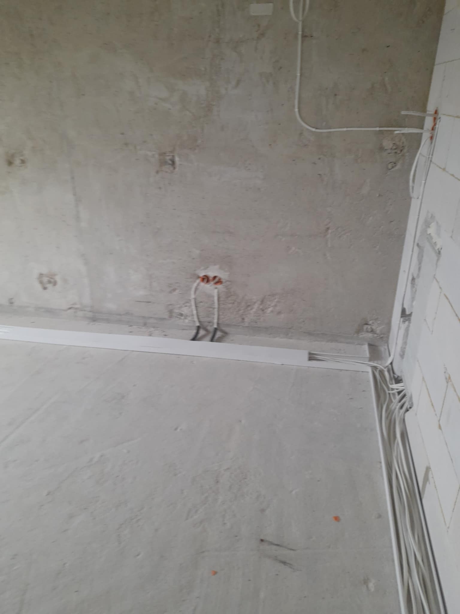 Surowy stan: wystające kable elektryczne ze ściany w nowym budynku, białe korytka kablowe wzdłuż ścian, szare ściany w tle. Instalacja elektryczna w trakcie realizacji.