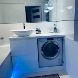 Biała łazienka z pralką w zabudowie pod umywalką, oświetlenie LED, lustro na ścianie. Nowoczesny design, funkcjonalne rozwiązanie.