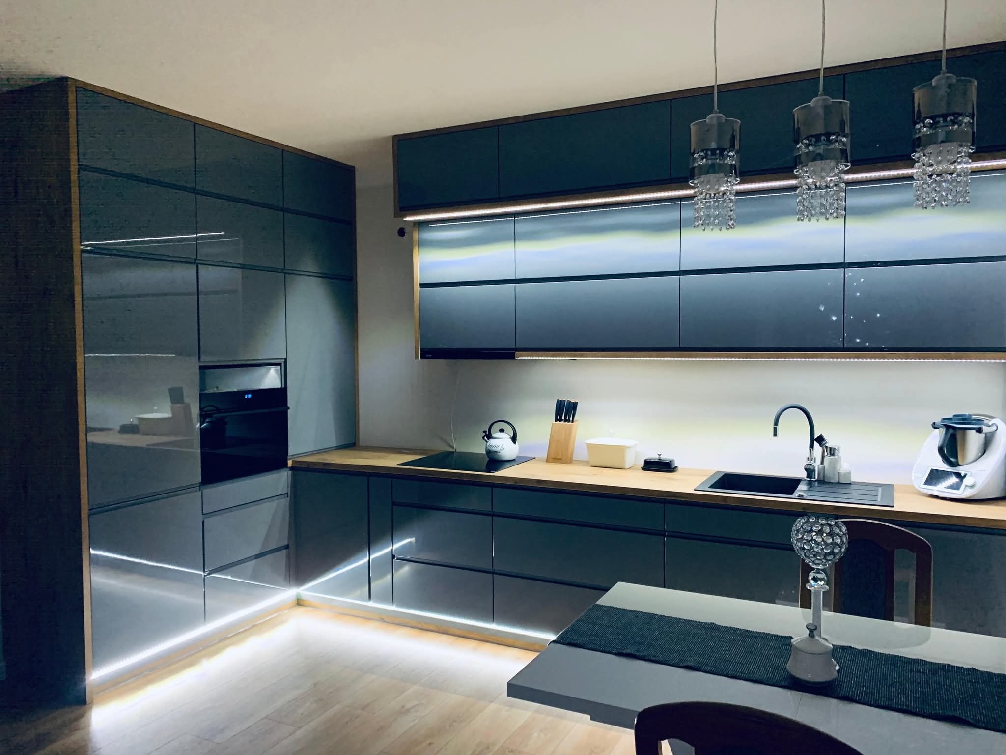Elegancka kuchnia z szarymi, lakierowanymi frontami, drewnianym blatem i subtelnym podświetleniem LED. Nad wyspą kuchenną wiszą ozdobne, kryształowe lampy.