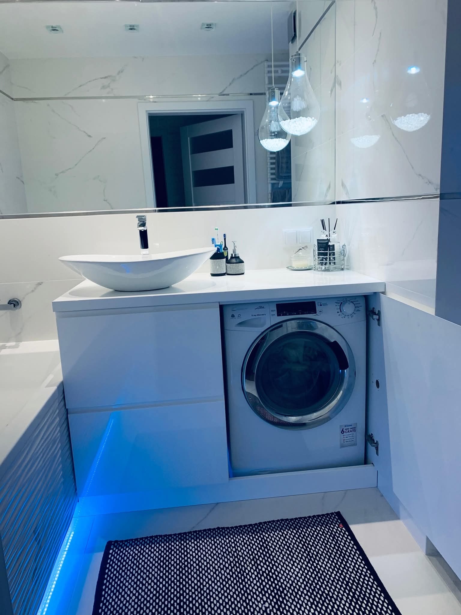 Biała łazienka z pralką w zabudowie pod umywalką, oświetlenie LED, lustro na ścianie. Nowoczesny design, funkcjonalne rozwiązanie.