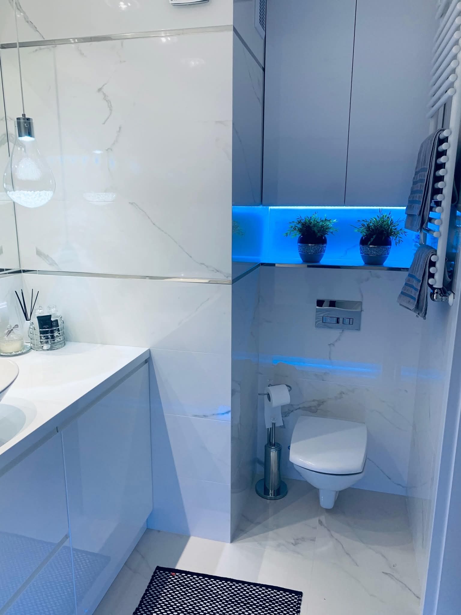 Nowoczesna, biała łazienka z marmurowymi płytkami, podświetlanymi półkami i wiszącą toaletą. Minimalistyczny design z akcentami roślinnymi i dekoracyjnym oświetleniem.