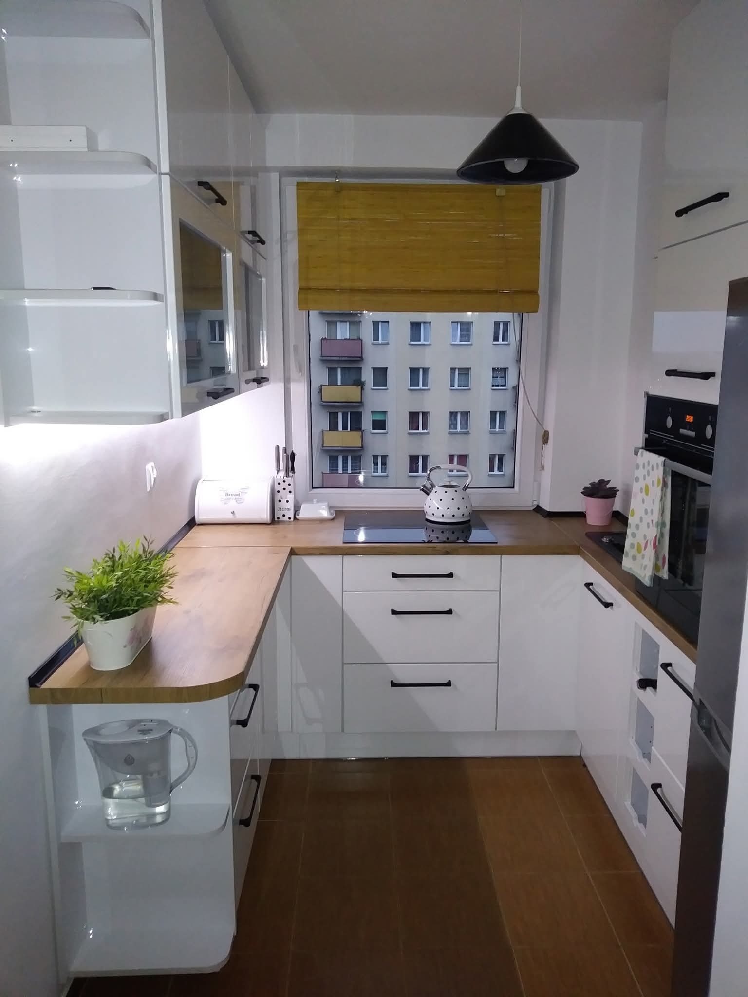 Nowoczesna, jasna kuchnia z drewnianym blatem i czarnymi uchwytami, widok na blok przez okno z bambusową roletą. Minimalistyczny design.