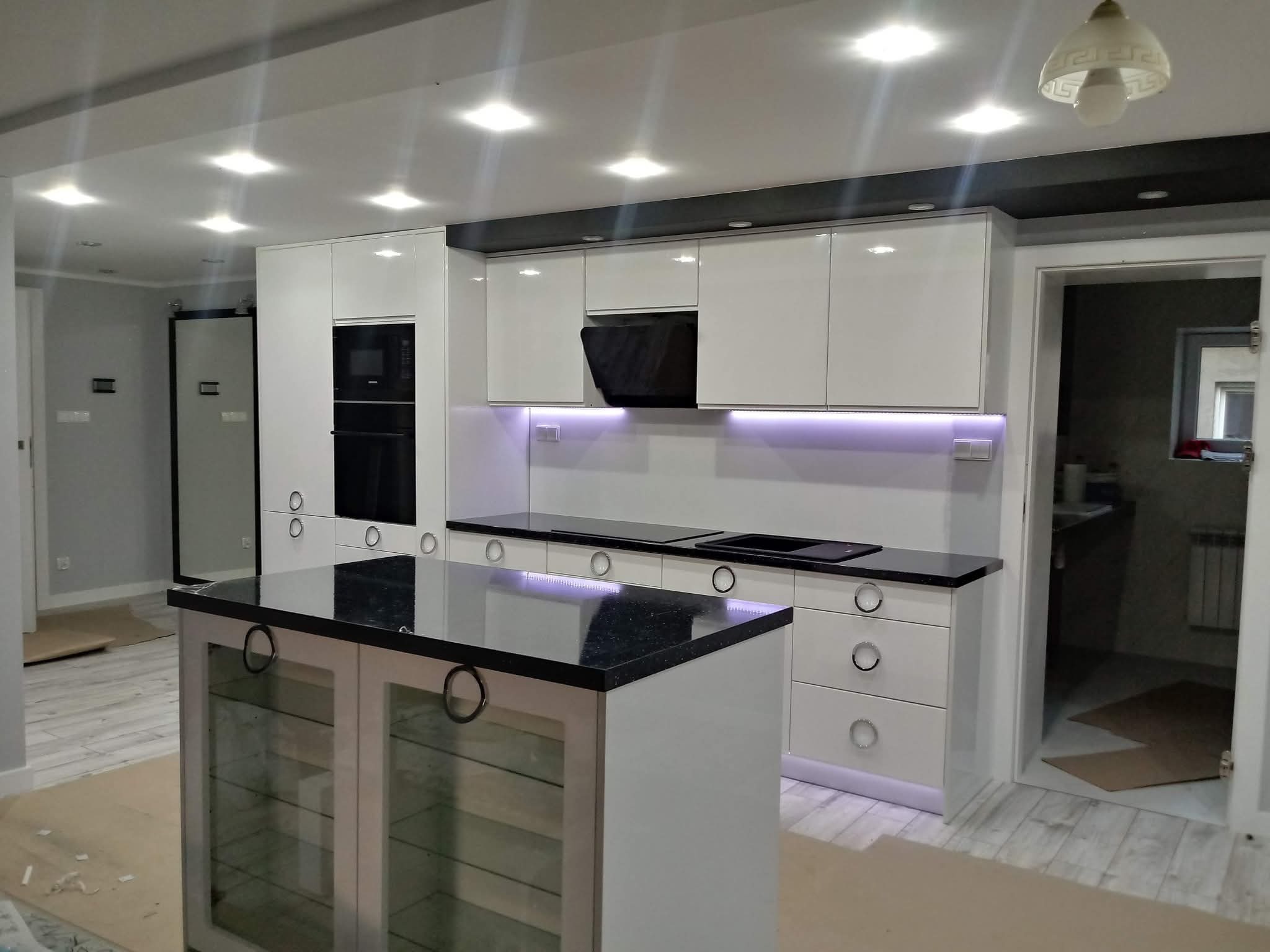 Biała kuchnia na wymiar z czarnym blatem i wyspą, oświetlenie LED pod szafkami. Nowoczesny design, szklane fronty wyspy.