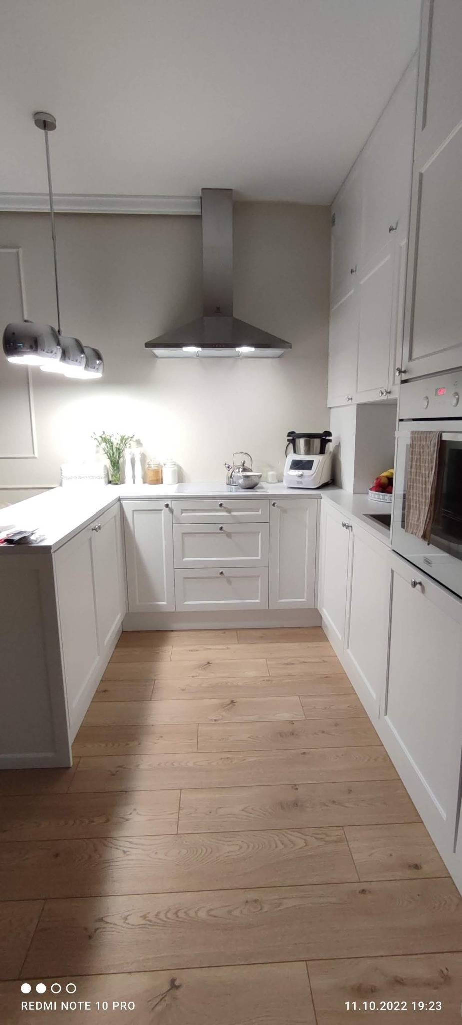 Jasna kuchnia w kształcie litery U z drewnianą podłogą i sprzętem AGD. Nad blatem wazon z kwiatami, nowoczesny okap. Uwagę przykuwają chromowane lampy.