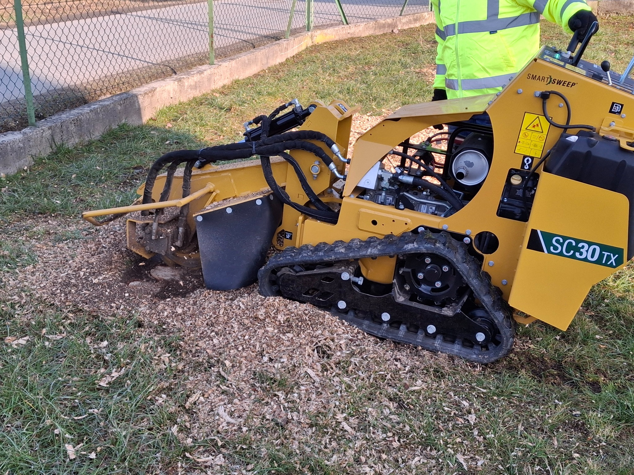 Żółta frezarka do pni SC30TX na gąsienicach, z widocznymi wiórami po frezowaniu, na trawniku. W tle ogrodzenie z siatki.