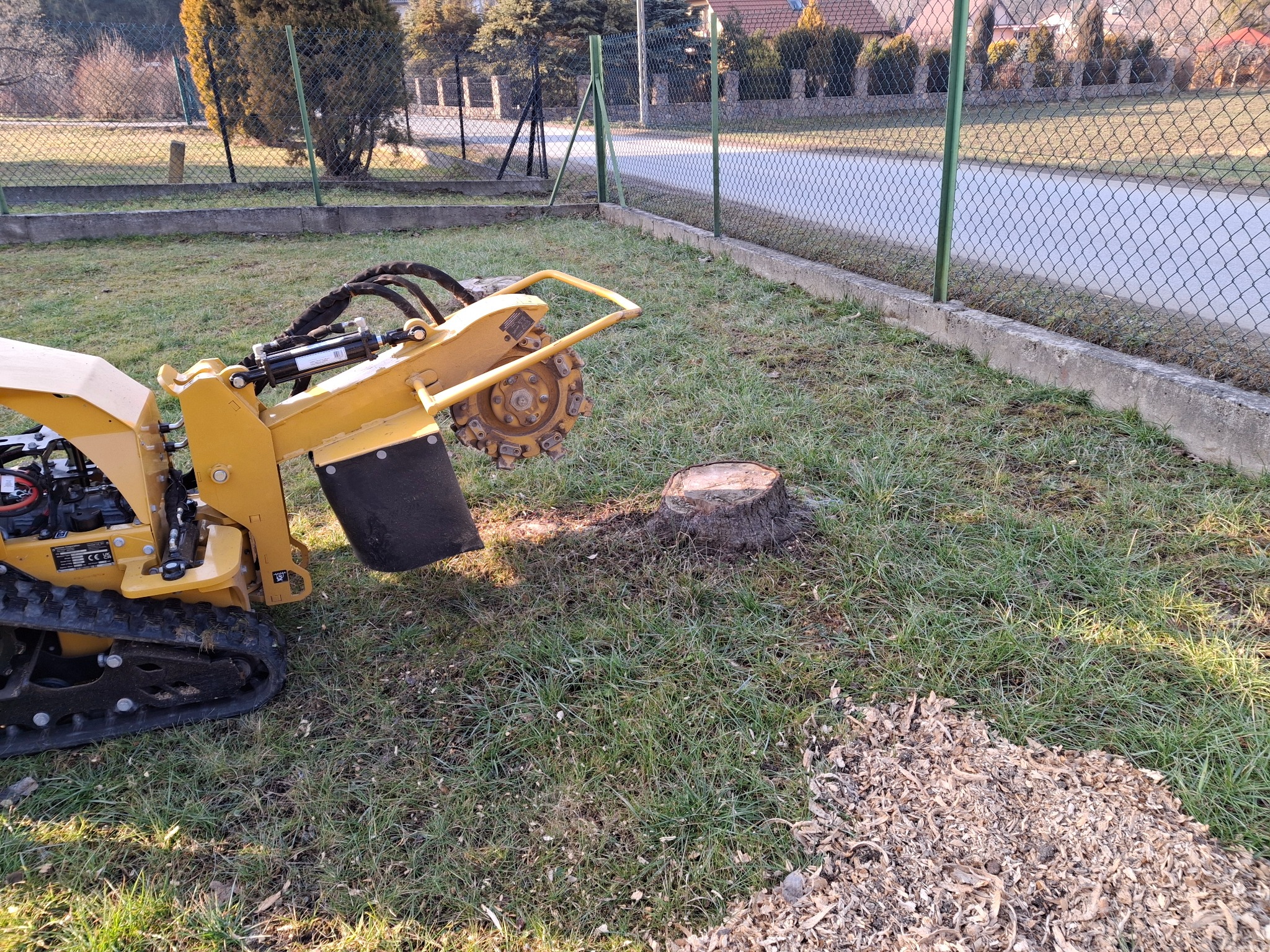 Żółta frezarka do pni drzew w ogrodzie, obok ścięty pień i zrębki. Widoczne ogrodzenie i domy w tle. Maszyna gotowa do pracy.