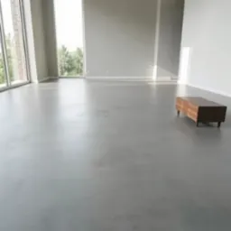 Szara, gładka posadzka w przestronnym, jasnym wnętrzu z dużymi oknami. Na podłodze stoi mała, drewniana szafka. Widok na zieleń za oknem.