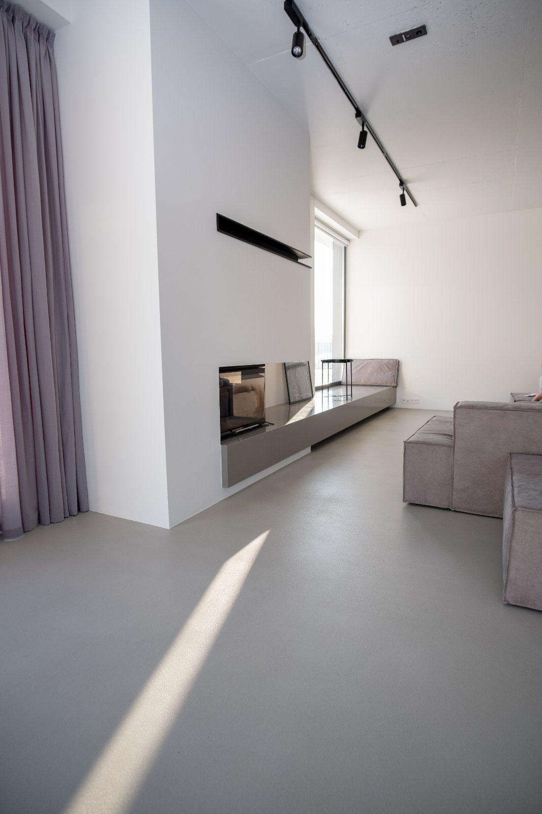 Minimalistyczne wnętrze z szarą posadzką z mikrocementu i wbudowanym kominkiem. Promień światła podkreśla gładką powierzchnię podłogi.