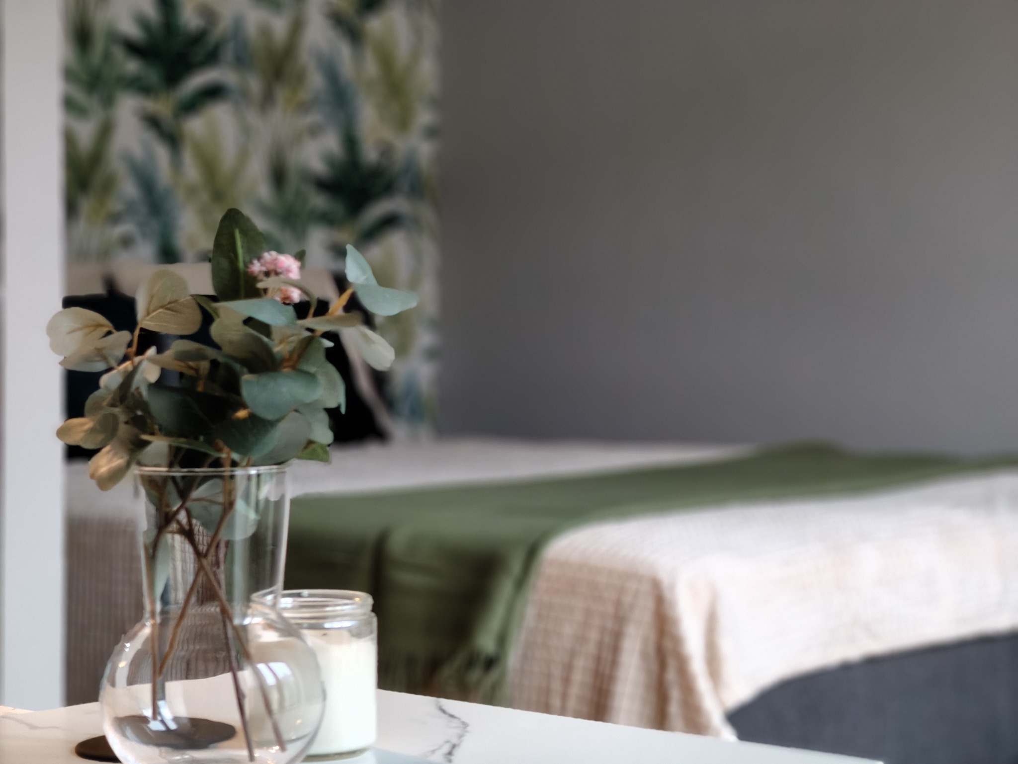 Jasne wnętrze sypialni z dekoracyjnym bukietem w szklanym wazonie na marmurowym blacie. W tle łóżko z zielonym pledem i tapeta w liście.