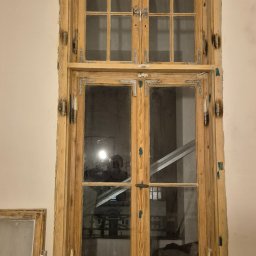 Serwis i naprawa okien Jakub Jewuła - Drewniane okno ze szprosami, w trakcie renowacji, z odsłoniętym usłojeniem drewna i starymi okuciami. W szybie widoczne odbicie osoby z telefonem.
