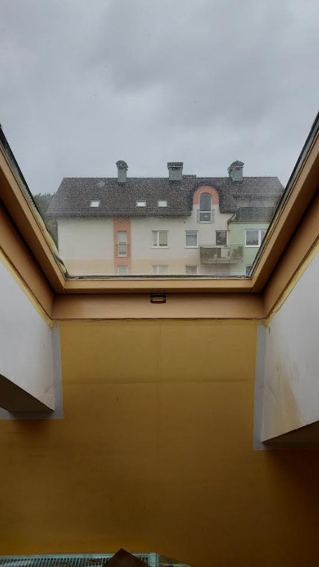 Zamknięte okno dachowe z widokiem na budynek mieszkalny w pochmurny dzień. Widoczne elementy konstrukcyjne okna i żółta ściana wewnętrzna.