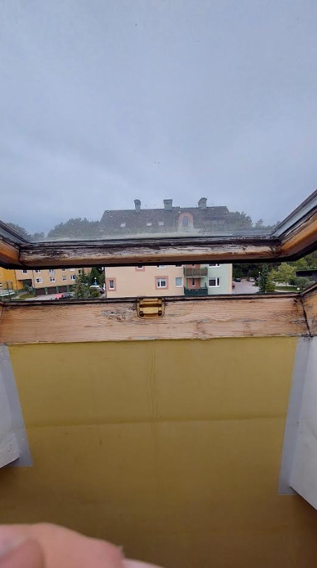 Otworzone okno dachowe z widocznymi śladami zużycia i budynku w tle. Widoczny mechanizm otwierania okna.