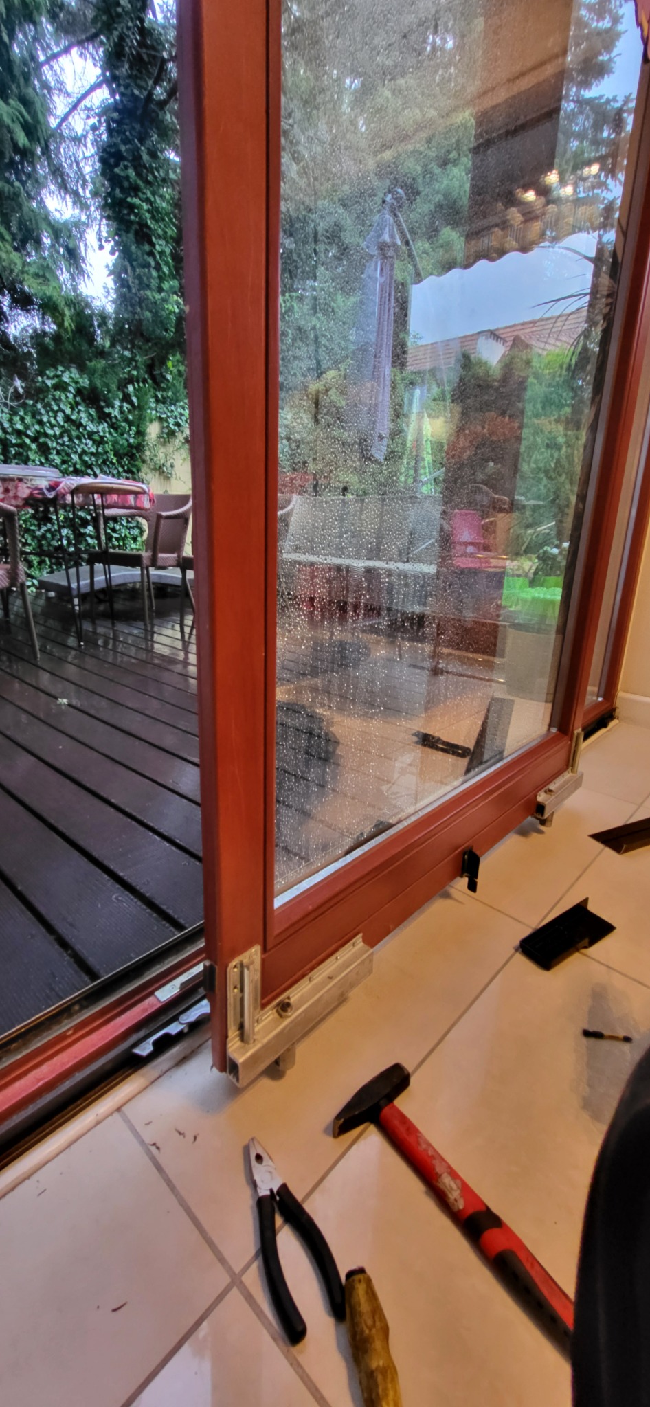 Demontaż brązowej ramy okiennej z widocznymi okuciami i narzędziami na podłodze. W tle deski tarasu i zieleń.