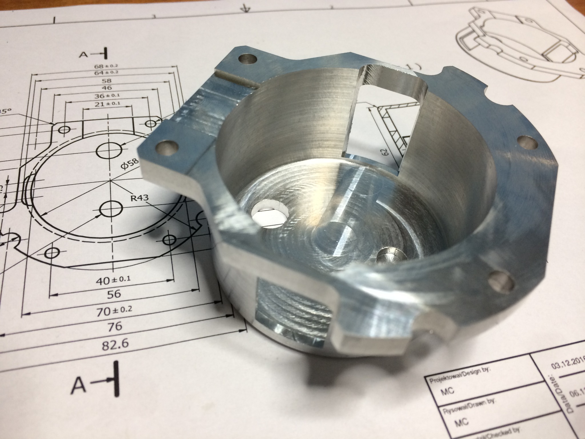 Metalowy element z otworem, obok rysunek techniczny z wymiarami. Widoczne ślady obróbki. Detal wykonany z precyzją, na rysunku daty i oznaczenia.