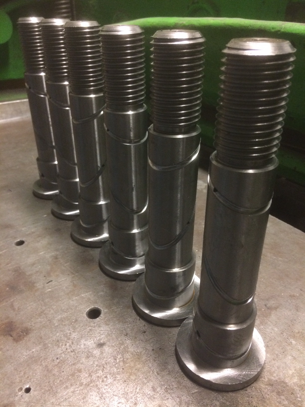 Srebrne, metalowe śruby z gwintem ułożone w rzędzie na metalowym stole warsztatowym. Widoczne ślady obróbki na powierzchni śrub.