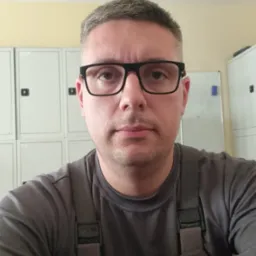 Portret mężczyzny w okularach i roboczym kombinezonie na tle metalowych szafek i białej tablicy. Ujęcie z bliska, neutralny wyraz twarzy.