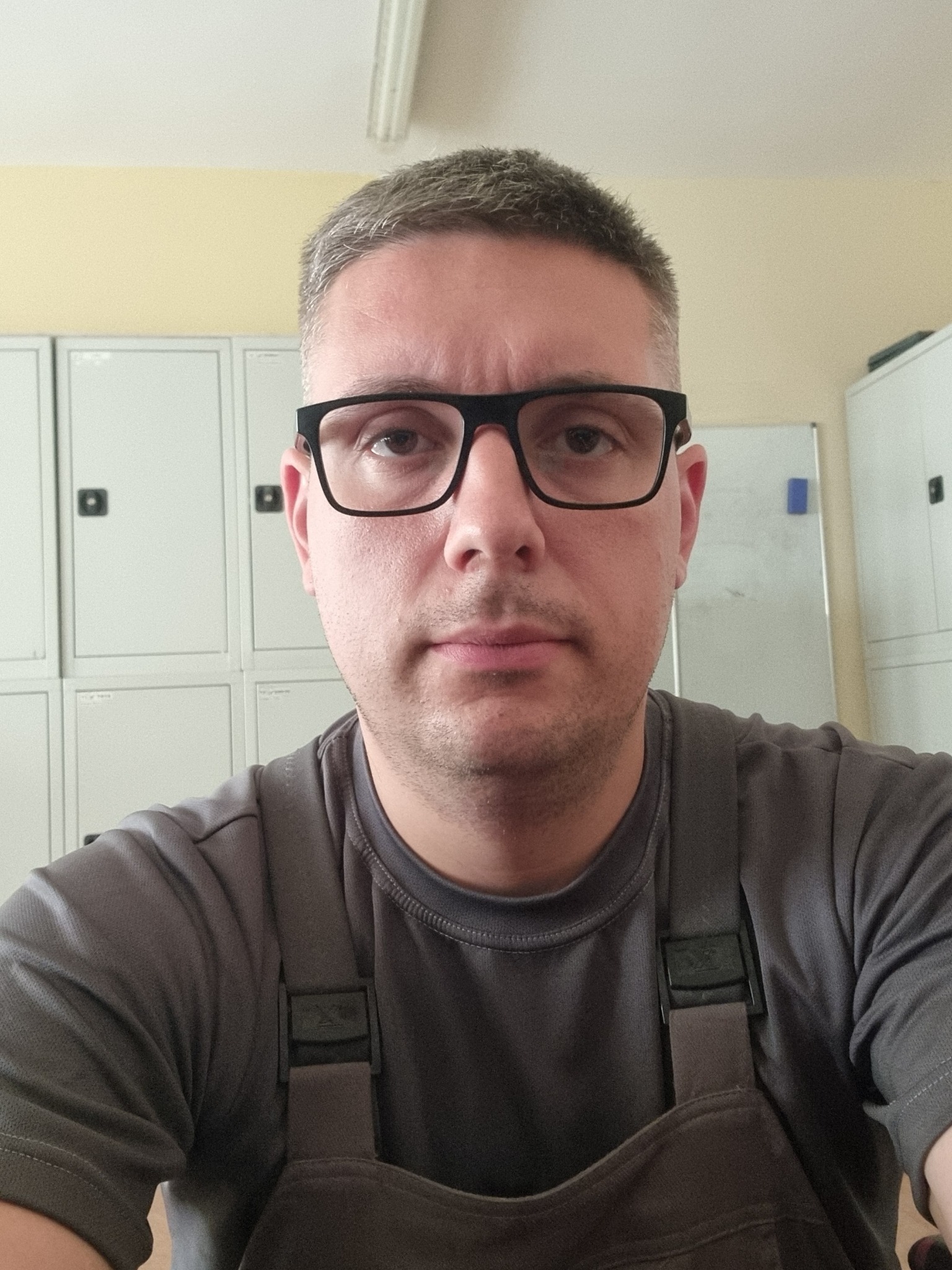 Portret mężczyzny w okularach i roboczym kombinezonie na tle metalowych szafek i białej tablicy. Ujęcie z bliska, neutralny wyraz twarzy.