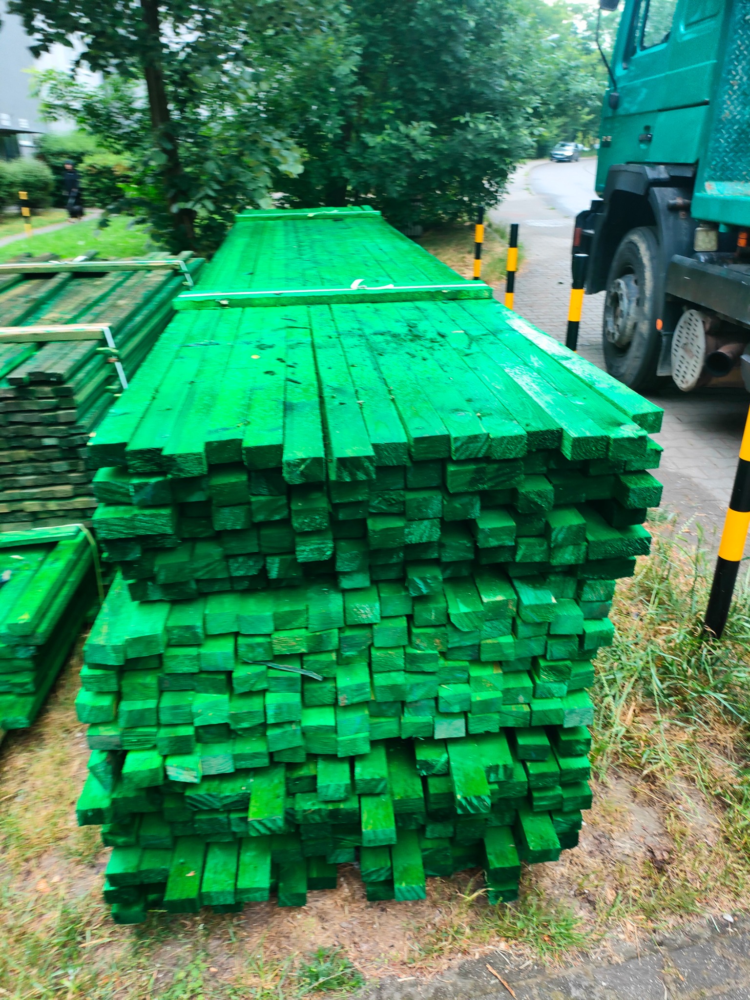 Stos zielonej tarcicy budowlanej ułożony obok zaparkowanej ciężarówki na tle zieleni. Elementy drewna zabezpieczone przed warunkami atmosferycznymi.