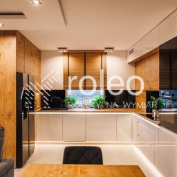 ROLEO - Drewniane żaluzje poziome w oknie nowoczesnej kuchni z drewnianymi i białymi szafkami. Widok na zieleń za oknem. Ciepłe oświetlenie wnętrza.