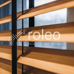 ROLEO - Drewniane lamele żaluzji wewnętrznych w ciepłym odcieniu, z czarnymi sznurkami, widziane pod kątem, z logo firmy nałożonym na zdjęcie.