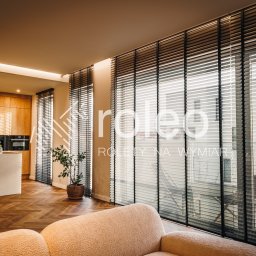 ROLEO - Eleganckie, czarne żaluzje wewnętrzne w luksusowym salonie z parkietem w jodełkę i nowoczesną kuchnią w tle. Widok na zewnątrz przez okno.