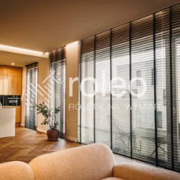 Eleganckie, czarne żaluzje wewnętrzne w luksusowym salonie z parkietem w jodełkę i nowoczesną kuchnią w tle. Widok na zewnątrz przez okno.