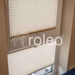 Beżowe plisy okienne zamontowane w oknie, widoczne od wewnątrz. Na parapecie dekoracyjne lampki. W tle budynek za oknem. U dołu kadru fragment niebieskiego fotela.