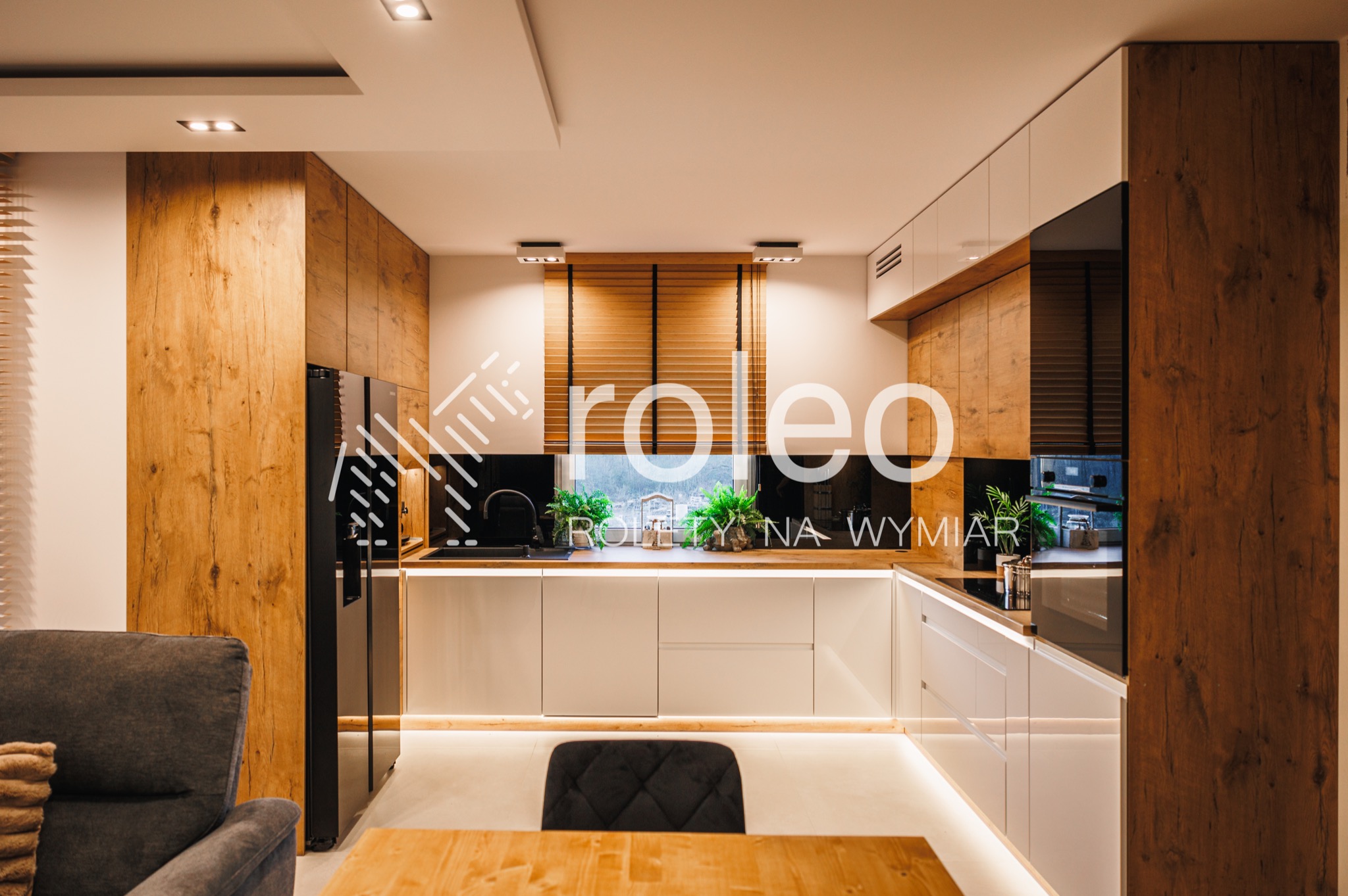 Drewniane żaluzje poziome w oknie nowoczesnej kuchni z drewnianymi i białymi szafkami. Widok na zieleń za oknem. Ciepłe oświetlenie wnętrza.