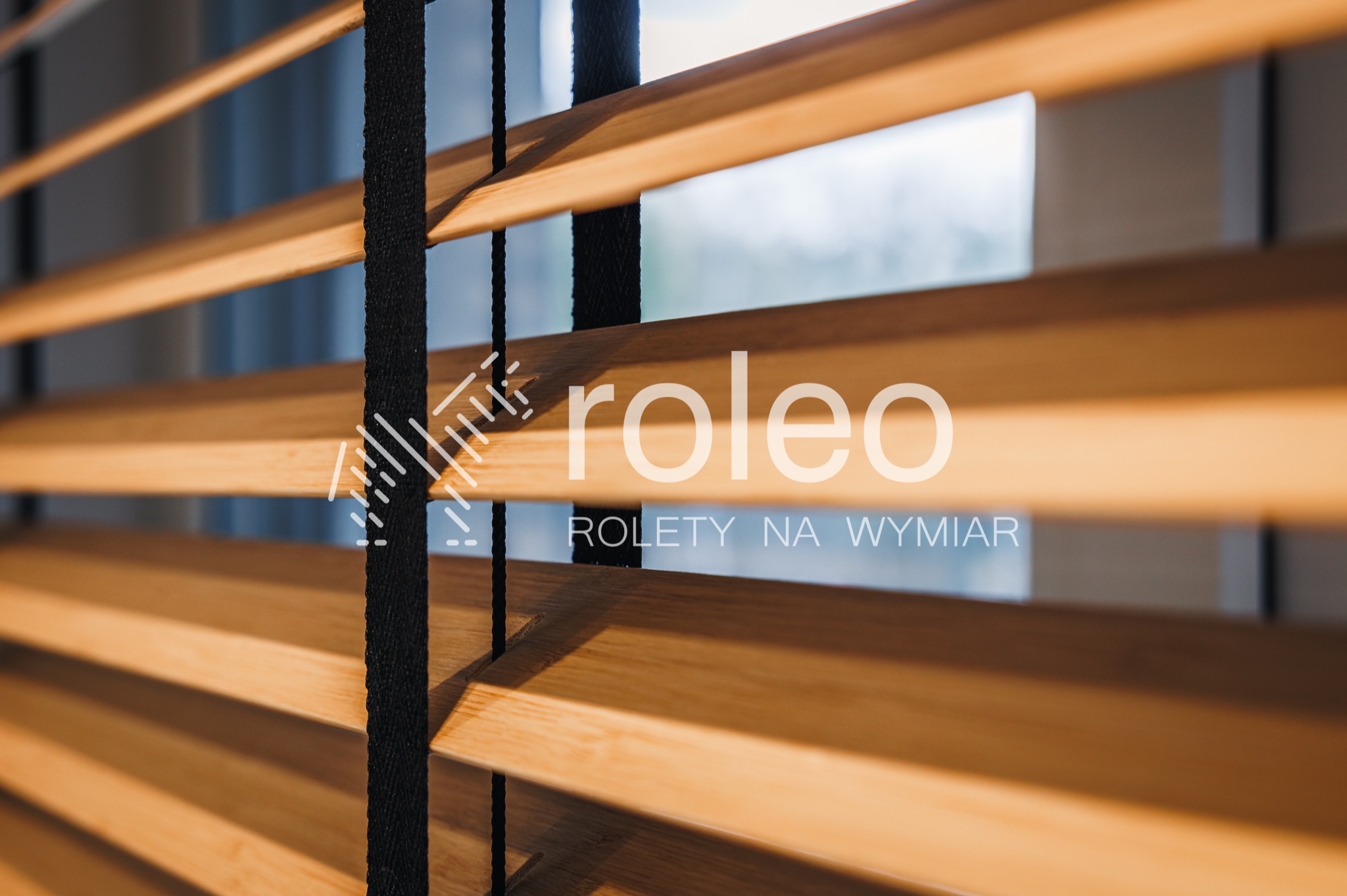 Drewniane lamele żaluzji wewnętrznych w ciepłym odcieniu, z czarnymi sznurkami, widziane pod kątem, z logo firmy nałożonym na zdjęcie.