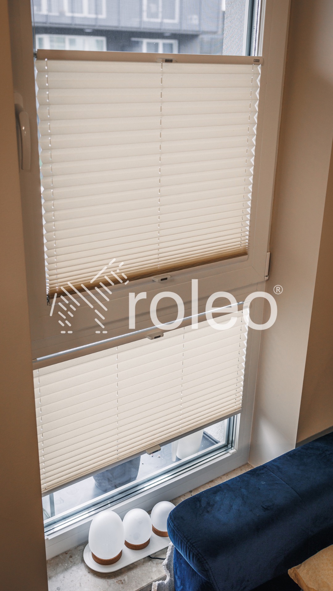 Beżowe plisy okienne zamontowane w oknie, widoczne od wewnątrz. Na parapecie dekoracyjne lampki. W tle budynek za oknem. U dołu kadru fragment niebieskiego fotela.