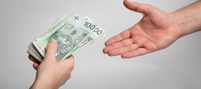 Dłoń wręcza plik banknotów 100 zł (PLN) drugiej dłoni na jasnym tle. Symboliczne przedstawienie transakcji finansowej, np. kredytu.