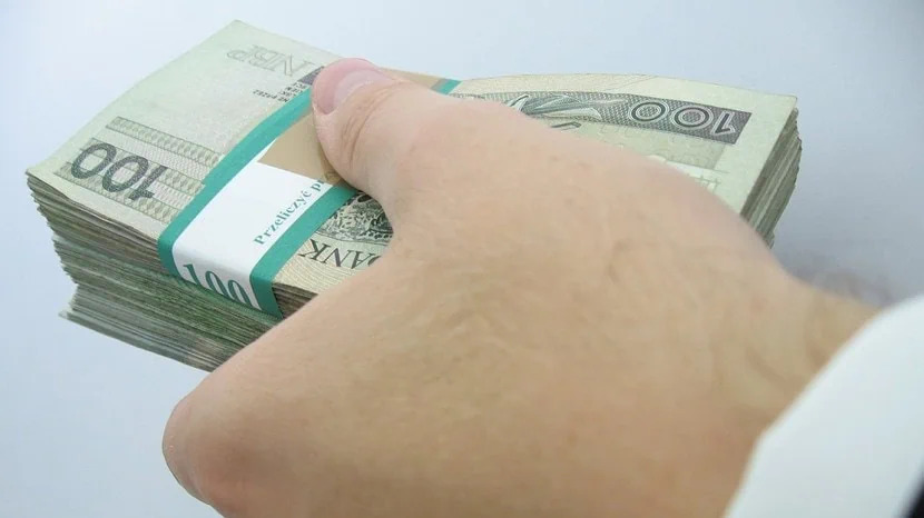 Dłoń trzymająca plik banknotów 100 PLN z banderolą NBP na jasnym tle, widoczny fragment białej koszuli.