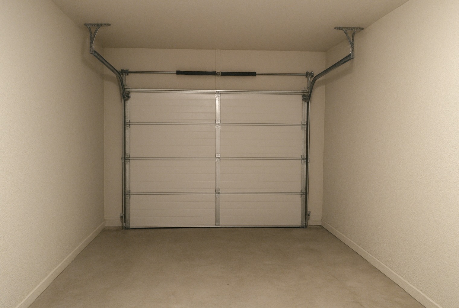 Wykończony garaż z jasnymi ścianami o delikatnej fakturze i betonową posadzką. Widoczna brama garażowa segmentowa, biała, z poziomymi przetłoczeniami.