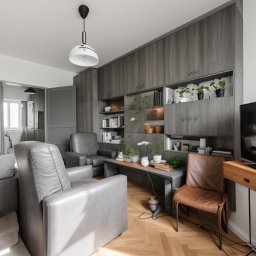 Kamil Dębkowski Nieruchomości - Nowoczesny salon z szarymi meblami, drewnianą podłogą w jodełkę i roślinami na półkach. Minimalistyczny design, stonowana kolorystyka, lampa wisząca.