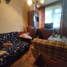 Kamil Dębkowski Nieruchomości - Umeblowane wnętrze pokoju z kanapą w renifery, brązową szafą i telewizorem przy oknie. Widoczne drewniane podłogi i lampa sufitowa.