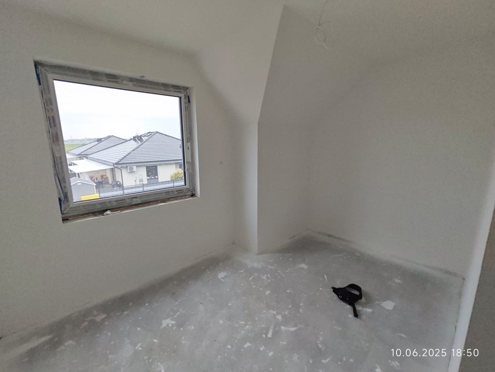 Poddasze w stanie deweloperskim z oknem, przez które widać domy. Na podłodze widoczne ślady po wylewce. W rogu leży narzędzie.