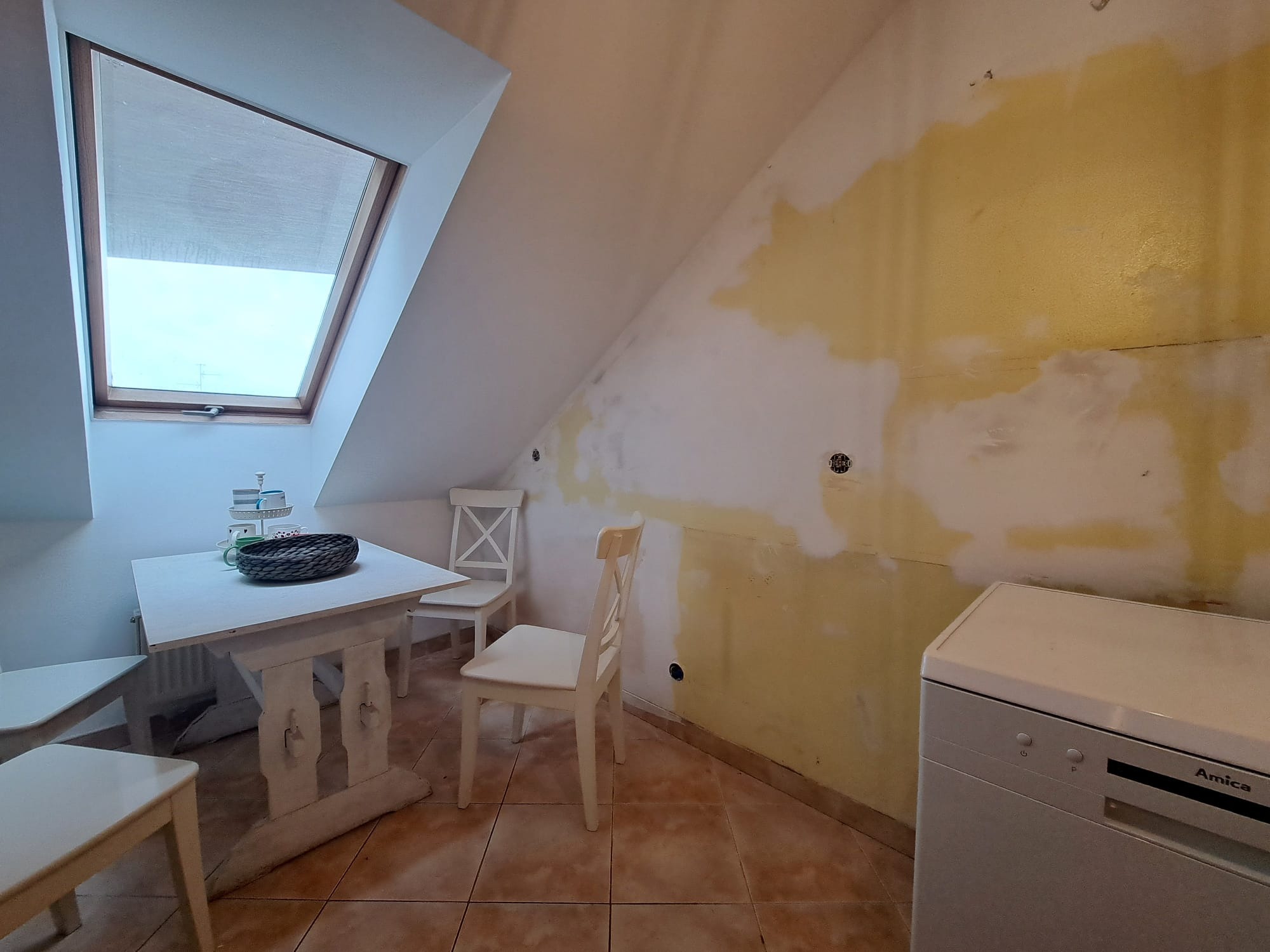 Poddasze w trakcie remontu: okno dachowe, białe meble kuchenne, ściana z plamami farby, widoczna pralka Amica. Etap przygotowania pomieszczenia do wykończenia.