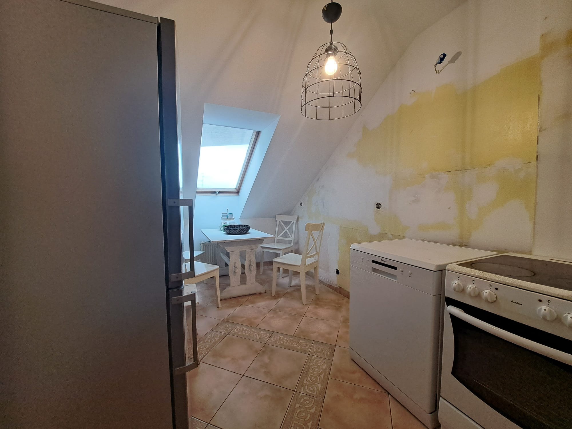 Klimatyczna kuchnia na poddaszu z oknem dachowym i białymi meblami. Ściany w trakcie remontu, widoczna instalacja elektryczna i fragment lodówki.