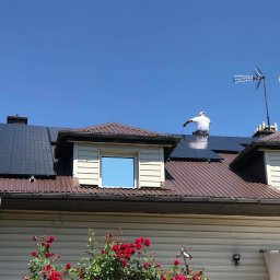 INVICTEL PIKUŁA SPÓŁKA JAWNA - Instalacja paneli słonecznych na brązowym dachu domu z anteną satelitarną i telewizyjną. Pracownik w białej koszulce łączy przewody na panelach.