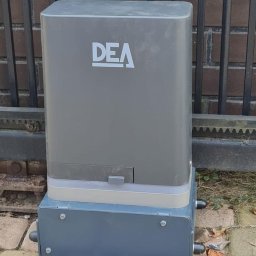 INVICTEL PIKUŁA SPÓŁKA JAWNA - Szary automat do bram DEA zamontowany na kostce brukowej przy metalowym ogrodzeniu z ceglanym murem w tle. Widoczne śruby mocujące i przewody.