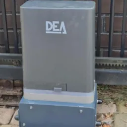 Szary automat do bram DEA zamontowany na kostce brukowej przy metalowym ogrodzeniu z ceglanym murem w tle. Widoczne śruby mocujące i przewody.