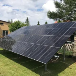 Panele fotowoltaiczne zamontowane na metalowej konstrukcji na trawniku, z budynkiem i drzewami w tle, w słoneczny dzień. Widok z bliska na gotową instalację.