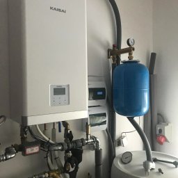 INVICTEL PIKUŁA SPÓŁKA JAWNA - Biały piec gazowy marki Kaisai z podłączeniami hydraulicznymi, zbiornikiem wyrównawczym i elementami instalacji w pomieszczeniu technicznym. Widoczne przewody i zawory.